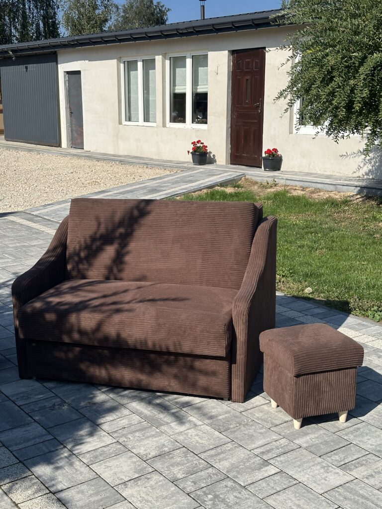 Brązowa dwuosobowa sofa z pasującą pufą, ustawiona na kostce brukowej przed domem